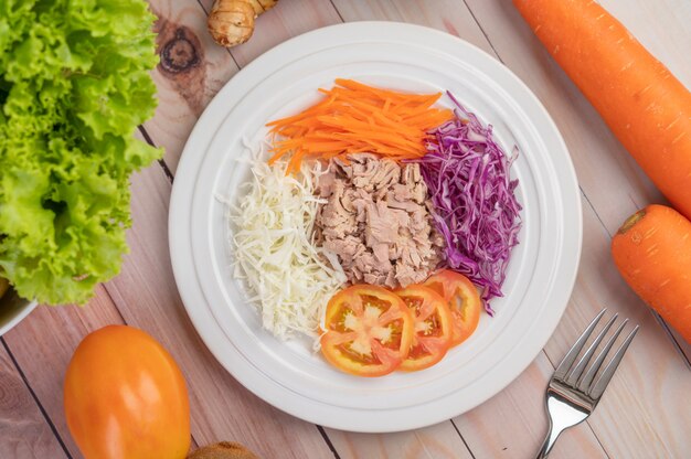 Салат из тунца с морковью, помидорами, капустой на белой тарелке на деревянном полу.