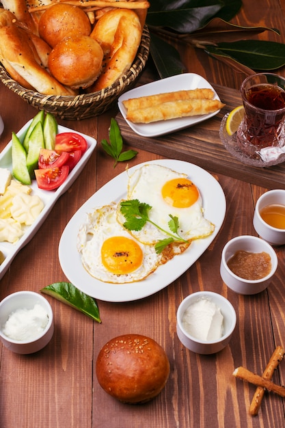 Free photo turkish breakfast with fried eggs, tomato, cucumber, cheese varieties, black green olives, honey, jam, cream cheese, galeta bread and glass of tea