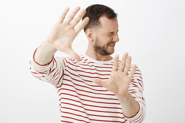 Turn light off. Portrait of displeased uncomfortable male model in casual outfit, pulling palms towards and turning face to hide from shiny light or punch