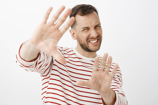 Turn off light it is to shiny. Displeased uncomfortable handsome adult man in striped outfit pulling hands towards to protect face, grimacing from discomfort