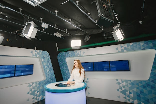 Tv present at studio preparing for new broadcasting. Smiling girl in white shirt sitting at table .