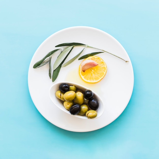 Twig, lemon slice, garlic clove and bowl of olives on plate over the blue background