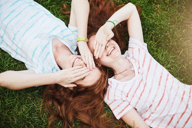 Twin sisters closing their eyes from sun, laying on ground on a summer day.