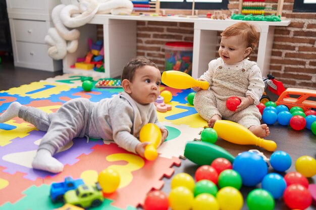 Two adorable babies playing with balls and bowling pin sitting on floor at kindergarten