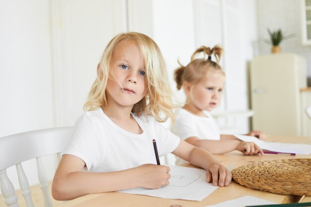 Two adorable Caucasian children doing homework together at wooden table. Cute seven year old boy with blond hair and blue eyes drawing at home with his little baby sister sitting