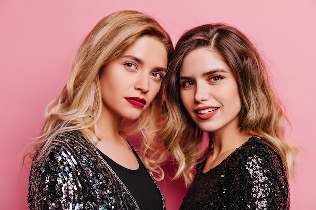 Two adorable girls posing with gently smile on pink wall. Pleased female models