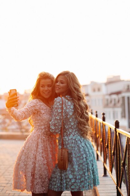 Two adorable girls smiling and posing in rose and blue flowery dresses taking self portrait at call phone Beautiful ladies with stylish straw bags making photo at smart phone together Sunset time