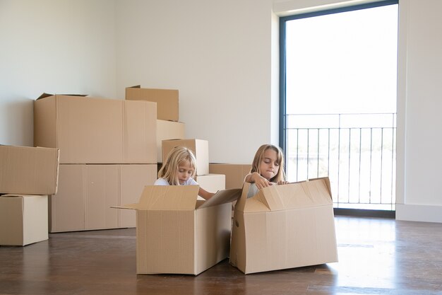 Two adorable girls unpacking things in new apartment, sitting on floor near open cartoon boxes