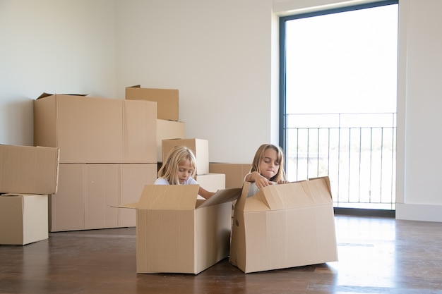 Two adorable girls unpacking things in new apartment, sitting on floor near open cartoon boxes
