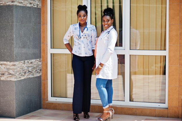 Two african american doctors female at lab coat with stethoscope posed outdoor against clinic