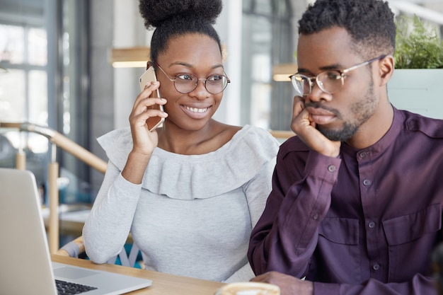 Two African American groupmates meet together for making project work or prepare for classes: happy dark skinned female speaks with friend via cell phone