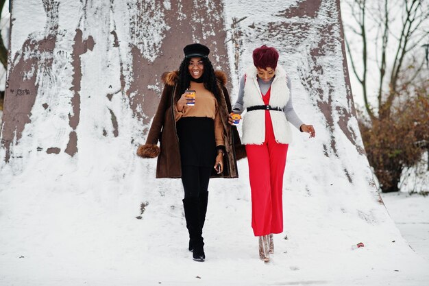 양가죽과 모피 코트를 입은 두 명의 아프리카계 미국인 여성이 겨울날 커피 한 잔과 함께 눈 덮인 배경에 대해 포즈를 취했습니다.