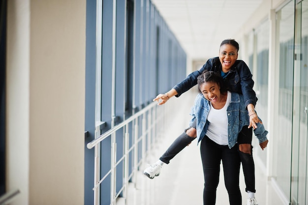 Two african woman friends in jeans jacket having fun jumped on shoulders indoor together