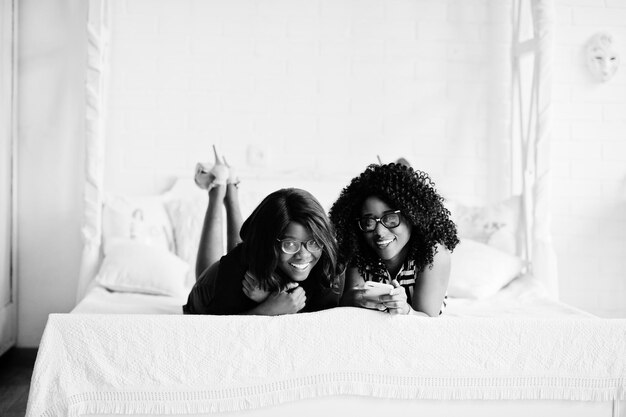 2人のアフリカの女性の友人がベッドの屋内の白い部屋に横たわって携帯電話を見ている眼鏡をかけています