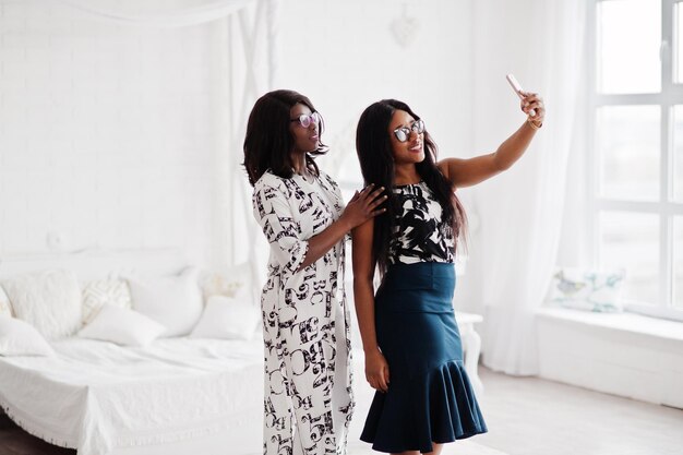 Two african woman friends wear on eyeglasses posed indoor white room and making selfie by cell phone