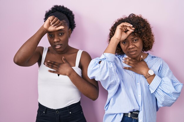 Two african women standing over pink background touching forehead for illness and fever flu and cold virus sick