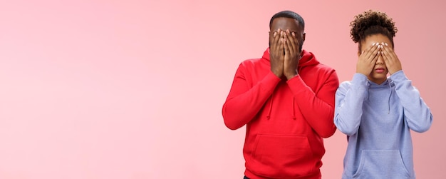 Two africanamerican man woman standing pink background hide faces close eyes palms tired looking lie