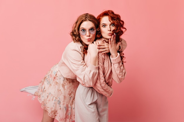 Two amazed caucasian ladies looking at camera. Best friends embracing on pink background.