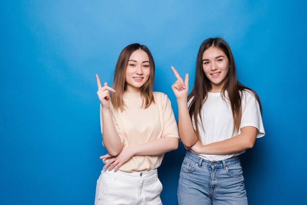 Two amazed mixed race women point with index fingers upwards, have happy expressions, isolated on blue wall.