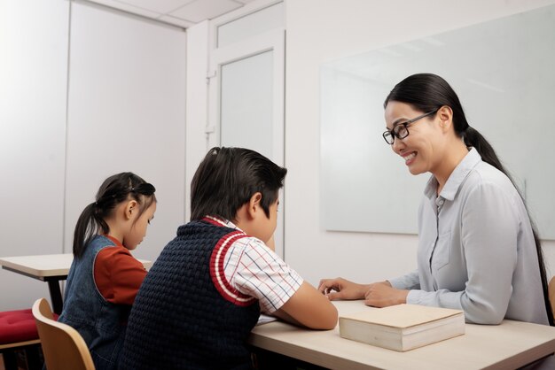 두 아시아 아이들이 교실에 앉아 소년과 이야기하는 안경에 교사 미소