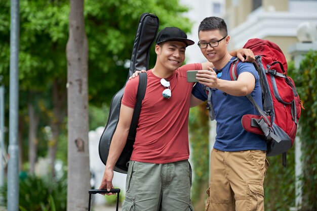 Two Asian male friends with luggage standing in city street, hugging and taking selfie