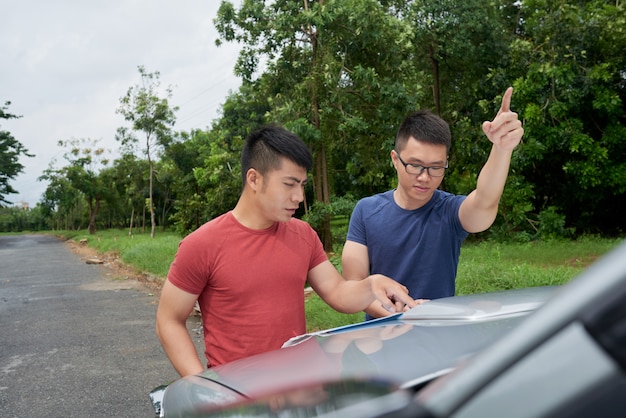 Two Asian men standing by car in road, looking at map and pointing forward