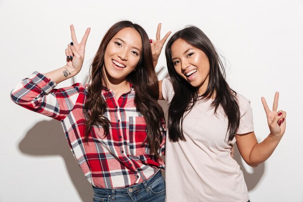 Two asian pretty cheerful ladies sisters showing peace gesture.