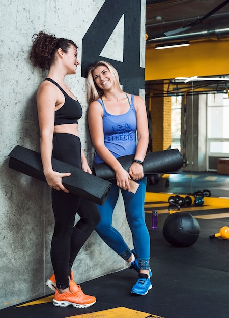 Две спортивные женщины с тренировочным матом, опираясь на стену в фитнес-клубе