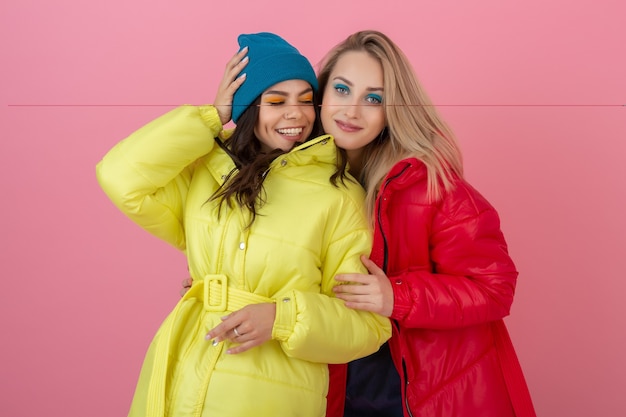 Two attractive active women posing on pink wall in colorful winter down jacket of bright red and yellow color, friends having fun together, warm coat fashion trend, crazy funny faces