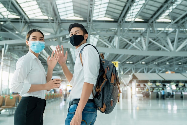 Two attractive asian businessman and businesswoman wear face mask protection hand gesture wave greeting and goodbye social distancing in departure airport terminal