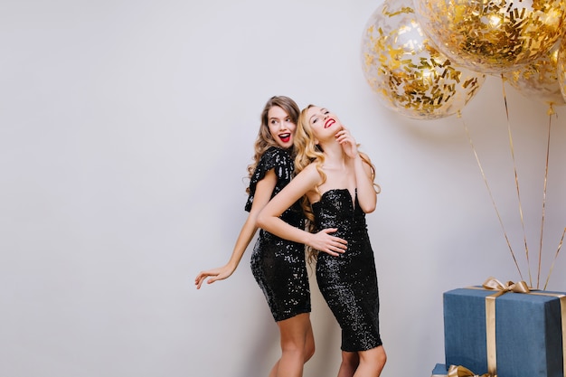 Две привлекательные модные молодые женщины в роскошных черных платьях празднуют вечеринку. Веселость, элегантный вид, улыбка, настоящие эмоции. Большой подарок, золотые шары, мишура.