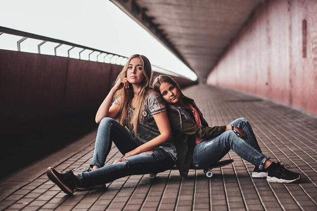 Two attractive friendly teenagers are sitting on the skateboard in long tunnel.