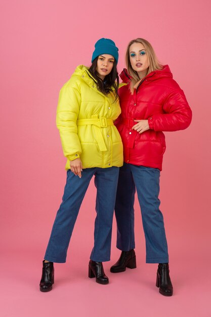 Two attractive girls posing on pink background in colorful winter down jacket of bright red and yellow color