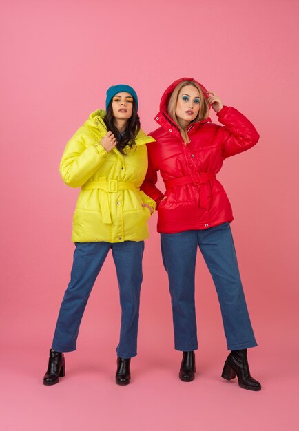 Two attractive girls posing on pink background in colorful winter down jacket of bright red and yellow color