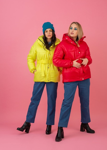 Two attractive girls posing on pink background in colorful winter down jacket of bright red and yellow color