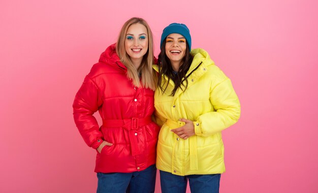 Two attractive girls posing on pink background in colorful winter down jacket of bright red and yellow color