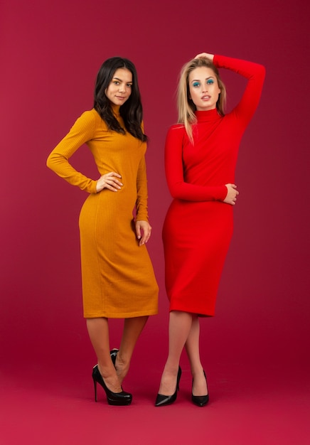 Two attractive stylish women in yellow and red autumn winter fashion knitted dress posing isolated  on red wall