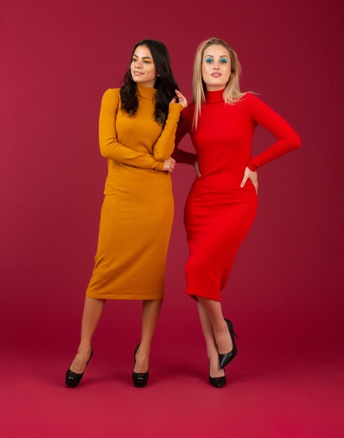 Two attractive stylish women in yellow and red autumn winter fashion knitted dress posing isolated  on red wall