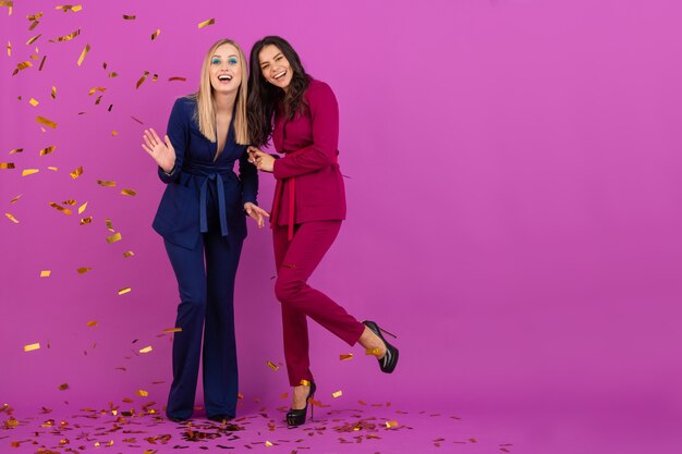 Two attractive women celebrating new year on violet wall in stylish colorful evening suits of purple and blue color, friends having fun together, fashion trend, golden confetti party mood