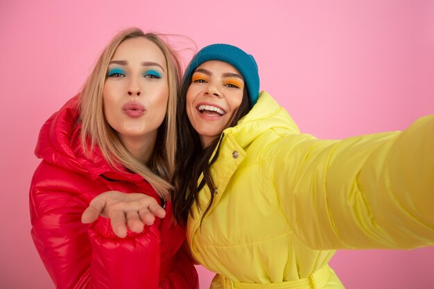 Two attractive women posing on pink background in colorful winter down jacket of bright red and yellow color, friends having fun together, warm clothes fashion trend, taking selfie