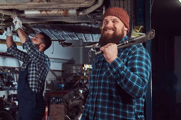 Two bearded brutal mechanics repair a car on a lift in the garage. Repairing car suspension in a service station.