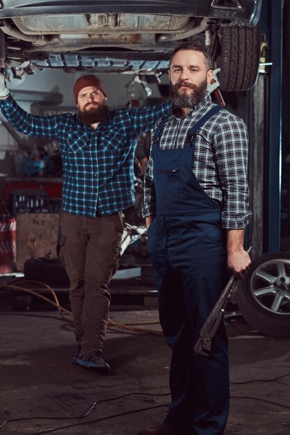 Два бородатых брутальных механика ремонтируют машину на подъемнике в гараже.