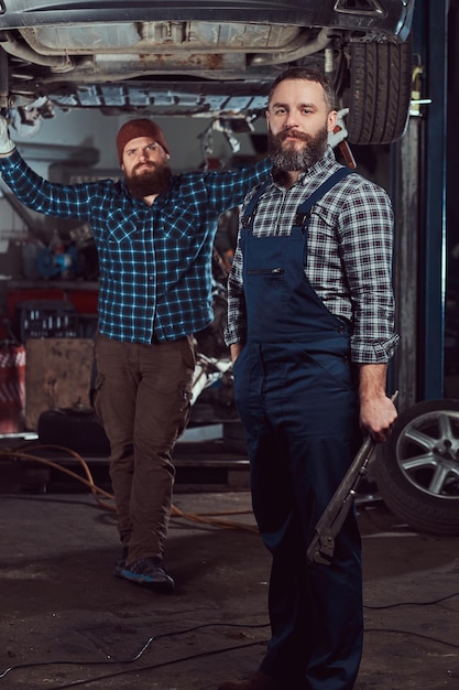 Два бородатых брутальных механика ремонтируют машину на подъемнике в гараже.