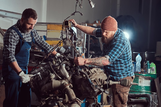 Два бородатых механика ремонтируют двигатель автомобиля, который поднимается на гидравлическом подъемнике в гараже. Станция обслуживания.