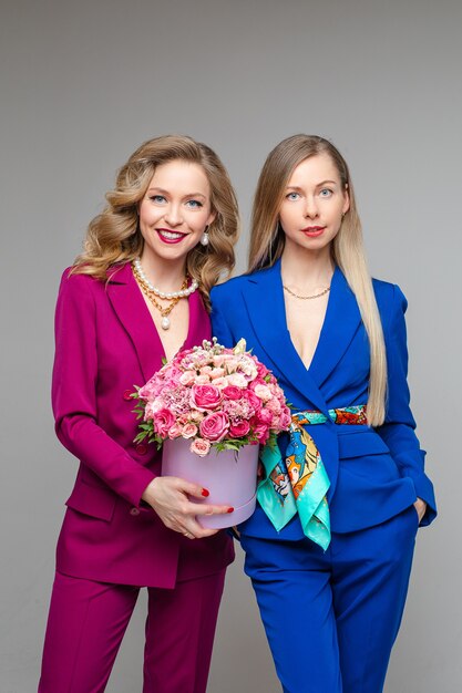 Two beautiful Caucasian blonde women with make up wearing stylish magenta and blue suits with jackets and trousers smiling at camera. Girl on the left holding beautiful flowers in hat box.