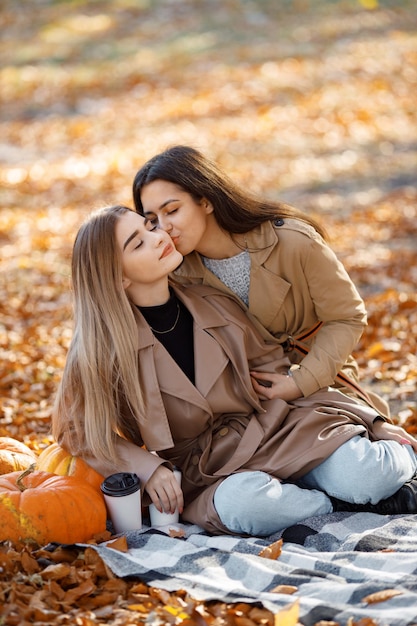 Two beautiful female friends spending time on a picnic blanket on the grass. Two young smiling sisters making picnic and hugging in autumn park. Brunette and blonde girls wearing coats.