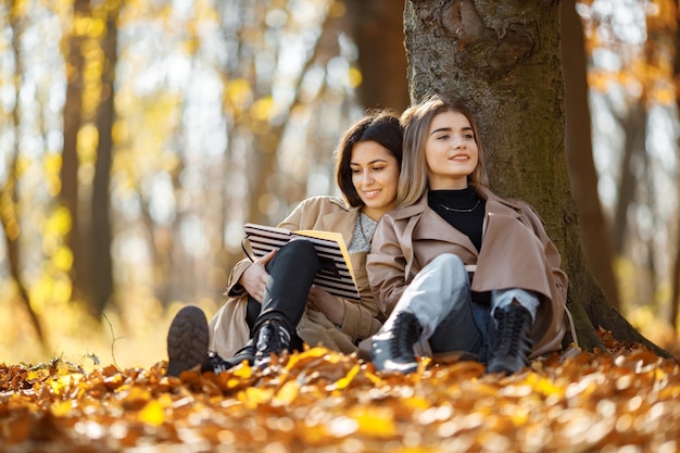 Two beautiful female friends spending time together. Two young smiling sisters sitting near tree and reading a book. Brunette and blonde girls wearing coats.