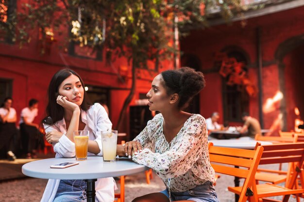 Two beautiful girls sitting at table with cocktails and cellphone dreamily talking while spending time together in cozy courtyard of cafe