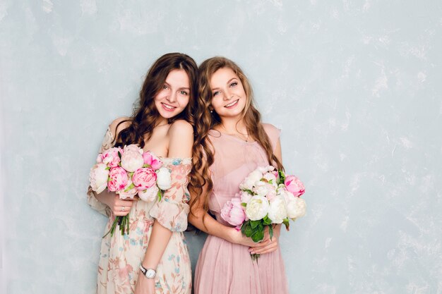 Две красивые девушки стоят в студии и держат букеты цветов.