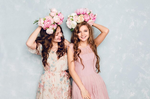 Two beautiful girls stand in a studio, play silly and have circlets of flowers on their heads. They wear light silk dresses. One is blond, and one is brunette. Both have curly hair.